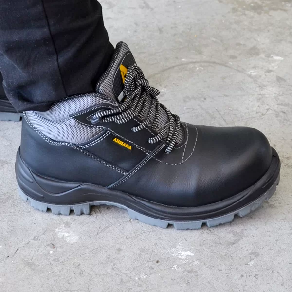 Un primer plano de una persona que lleva una resistente bota de trabajo negra con adornos grises y cordones sobre un piso de concreto. el maletero tiene una pequeña etiqueta amarilla con el texto "amblers" en el lateral.