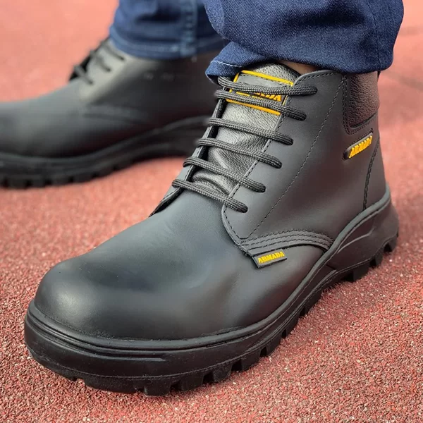 Un primer plano de una persona que lleva botas de cuero oscuro con costuras de color amarillo brillante y cordones negros. las botas se combinan con jeans azules enrollados, sobre una superficie rojiza.
