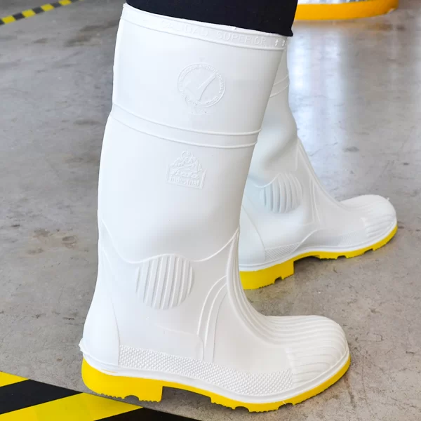 Un par de resistentes botas de goma blancas con suelas gruesas y amarillas, diseñadas específicamente para uso industrial, sobre un piso de concreto con líneas de seguridad amarillas. Las botas cuentan con logotipos en relieve y texto que enfatizan la calidad y la durabilidad.