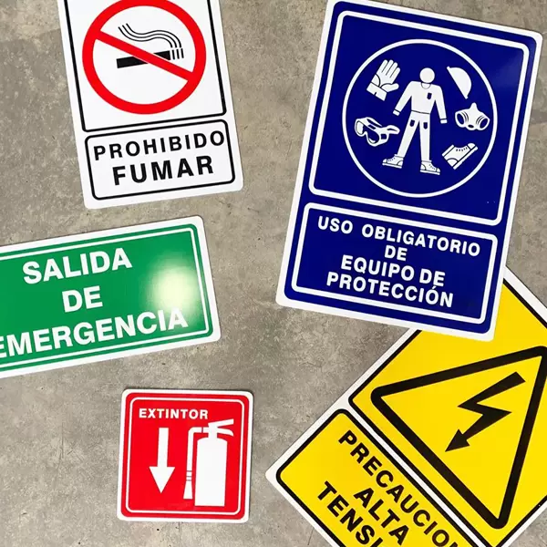 Cinco señales de seguridad sobre una superficie de concreto: "prohibido fumar" con un símbolo de no fumar, una señal azul de uso obligatorio del equipo de seguridad, una señal de salida de "salida de emergencia", una señal roja de extintor y una señal de precaución de alto voltaje.