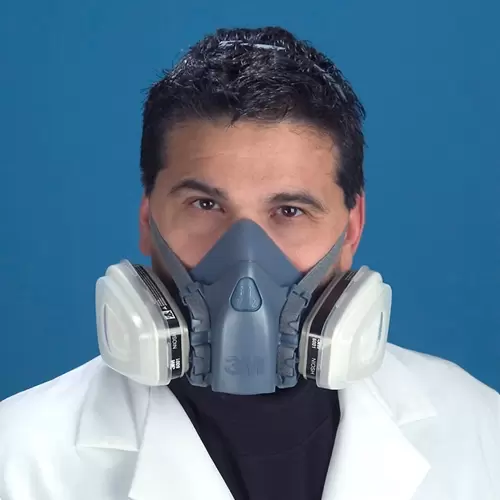 Un hombre que lleva una bata de laboratorio blanca y una máscara respiratoria gris de doble filtro le cubre la nariz y la boca, sobre un fondo azul. su cabello oscuro está ligeramente despeinado y mira directamente al espectador.