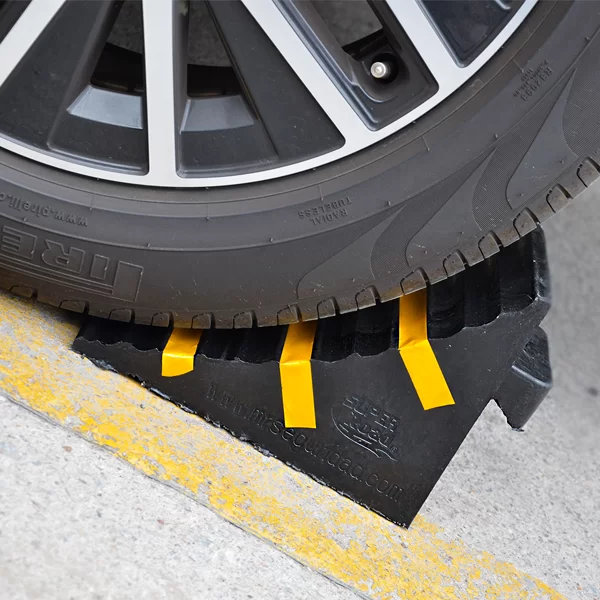 Un neumático de automóvil está parcialmente estacionado sobre un tope de goma negro con tiras reflectantes de color amarillo brillante, colocado sobre una superficie de concreto con una línea pintada de amarillo.