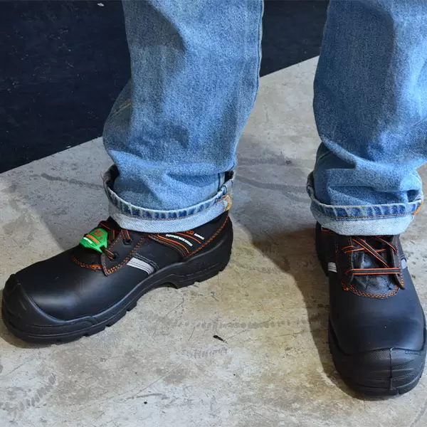 Una persona parada sobre una superficie negra, vestida con jeans azules y botas de trabajo negras con cordones y costuras de color naranja. un pequeño juguete verde colocado en el pie izquierdo añade un toque lúdico a la imagen.