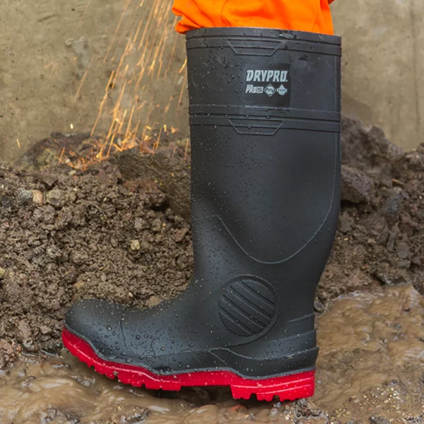 Un primer plano de una bota de goma negra con suela roja, con la etiqueta "drypak", parcialmente sumergida en agua fangosa y salpicada de tierra. Las chispas de una actividad cercana son visibles encima del maletero.