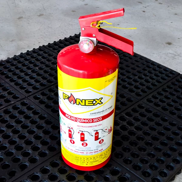 Un extintor rojo con una etiqueta amarilla que dice "fonex" en letras blancas e instrucciones en español, colocado sobre un piso de rejilla negra. el pasador de seguridad está intacto y el manómetro indica presión total.