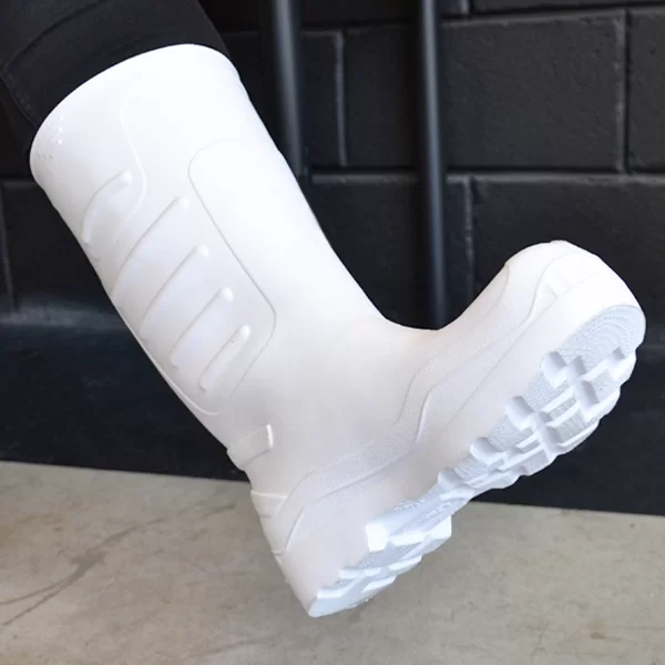 Una imagen en primer plano de una persona que lleva una bota blanca moderna con un diseño intrincado en la banda de rodadura, destacada contra una pared negra y un fondo de piso gris.